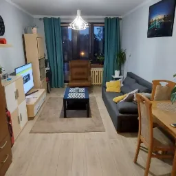 Widok na umeblowany salon z drewnianą podłogą, szarą sofą z żółtą poduszką, brązowym fotelem i czarno-białym stolikiem kawowym, oświetlony nowoczesną lampą wiszącą.