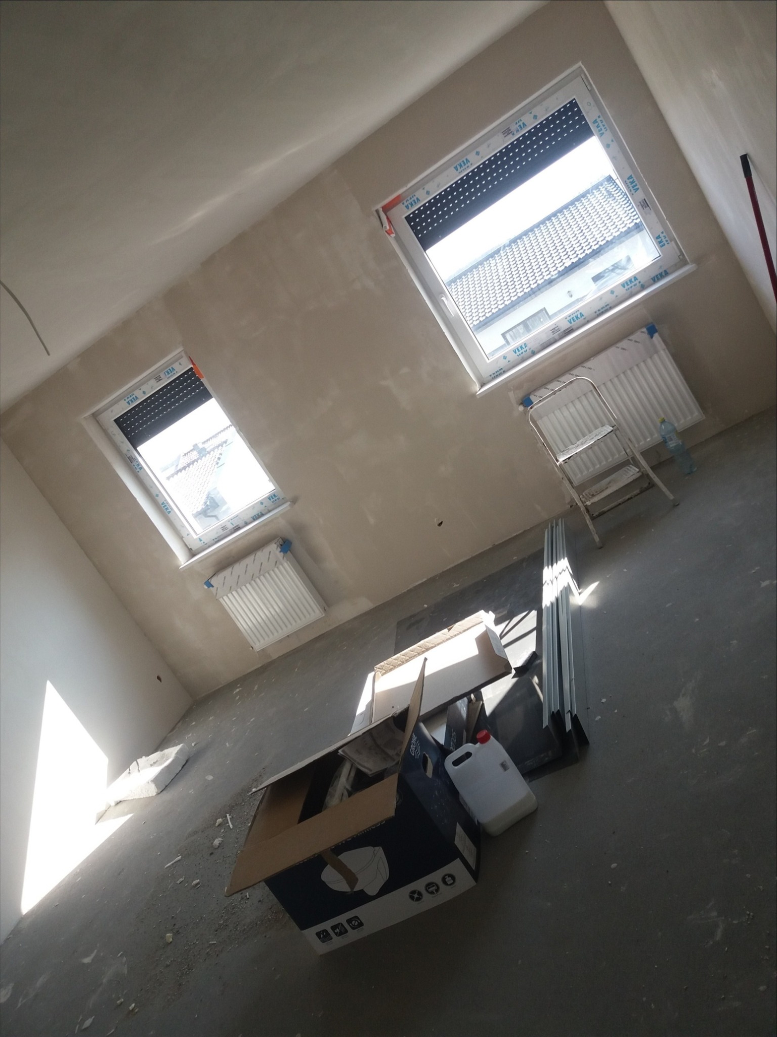 Wnętrze pokoju po tynkowaniu, widoczne dwa okna dachowe z roletami, grzejniki, drabina, karton i pojemnik z farbą, ściany w trakcie schnięcia tynku.