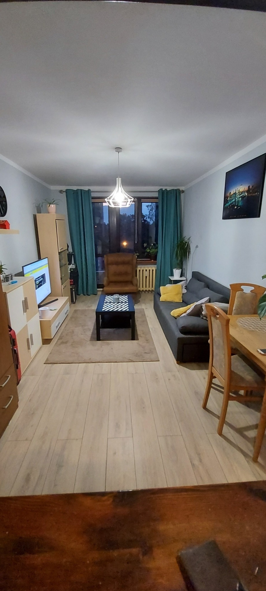 Widok na umeblowany salon z drewnianą podłogą, szarą sofą z żółtą poduszką, brązowym fotelem i czarno-białym stolikiem kawowym, oświetlony nowoczesną lampą wiszącą.