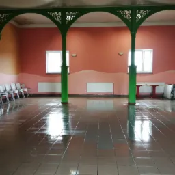 Sala z błyszczącą, mokrą podłogą z płytek, zielonymi kolumnami i łukami, krzesłami ustawionymi w rzędzie pod ścianą.