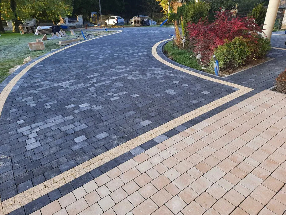 Szeroki podjazd z kostki brukowej w odcieniach szarości i beżu, oddzielony pasem żółtej kostki, otoczony zielenią i drzewami, z samochodami w tle.