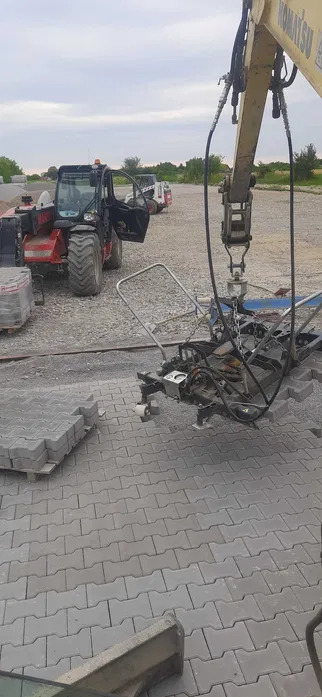 Układanie szarej kostki brukowej typu I z użyciem chwytaka podwieszonego do ramienia koparki Komatsu, w tle widoczny podnośnik teleskopowy Manitou i stos kostki brukowej na palecie.