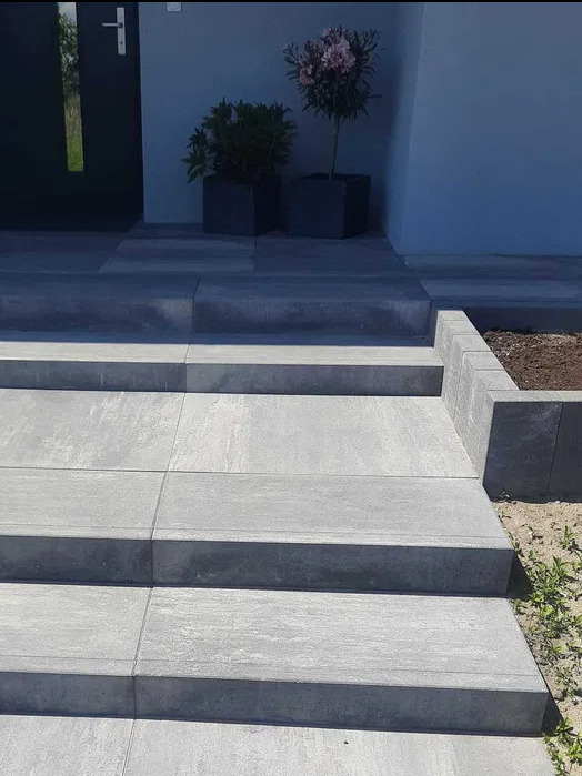 Nowoczesne schody wejściowe wyłożone szarą kostką brukową, prowadzące do drzwi wejściowych, z donicami z roślinami po bokach.