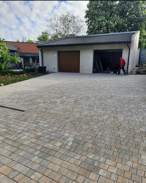 Ułożona kostka brukowa przed garażem z dwoma bramami, widoczny fragment ogrodu z kwiatami i przechodzący mężczyzna w czerwonej bluzie.