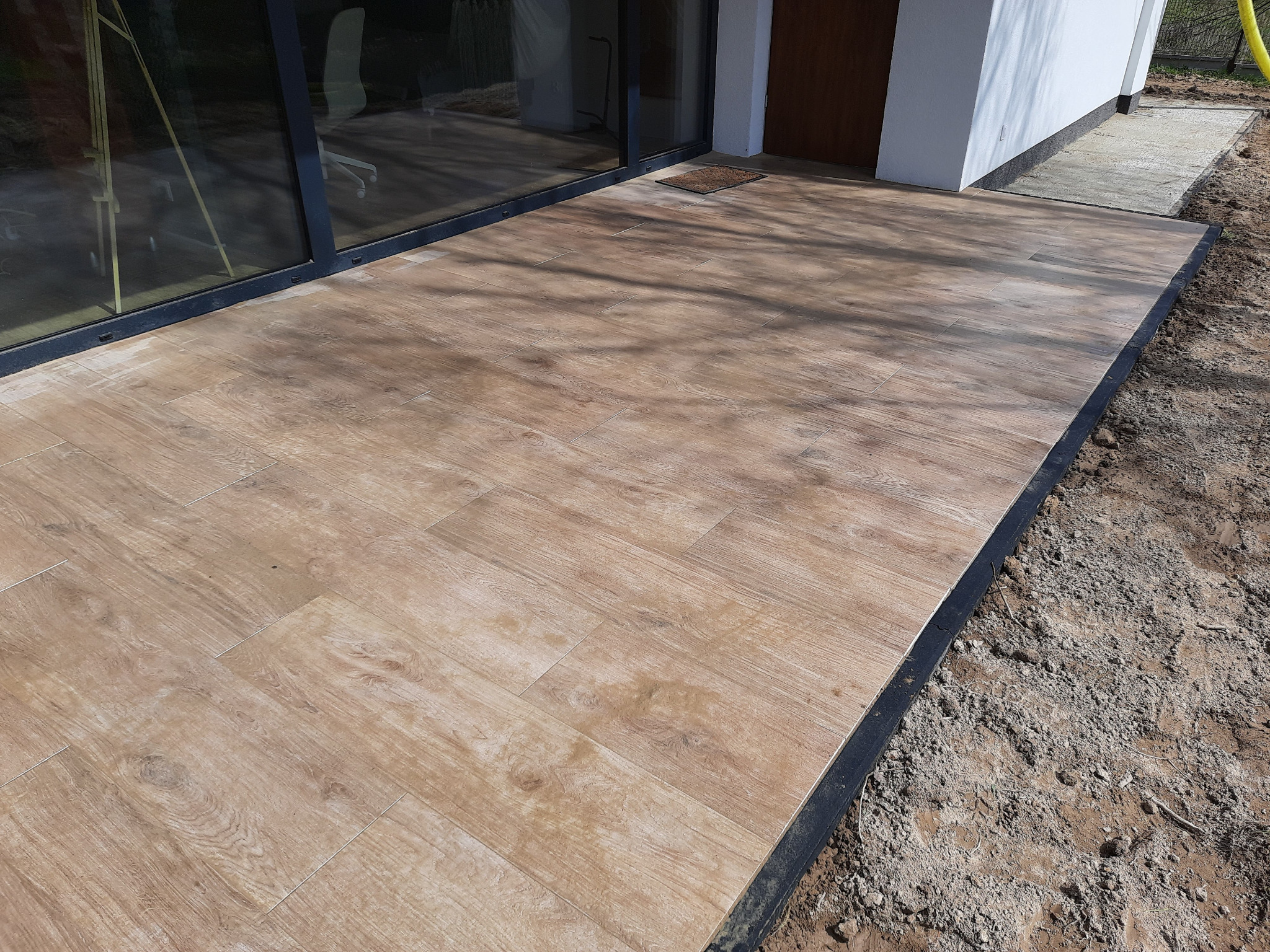 Jasne płytki imitujące drewno ułożone na tarasie przylegającym do domu z dużymi przesuwnymi drzwiami, widoczny fragment ogrodu z ziemią i fragment chodnika.