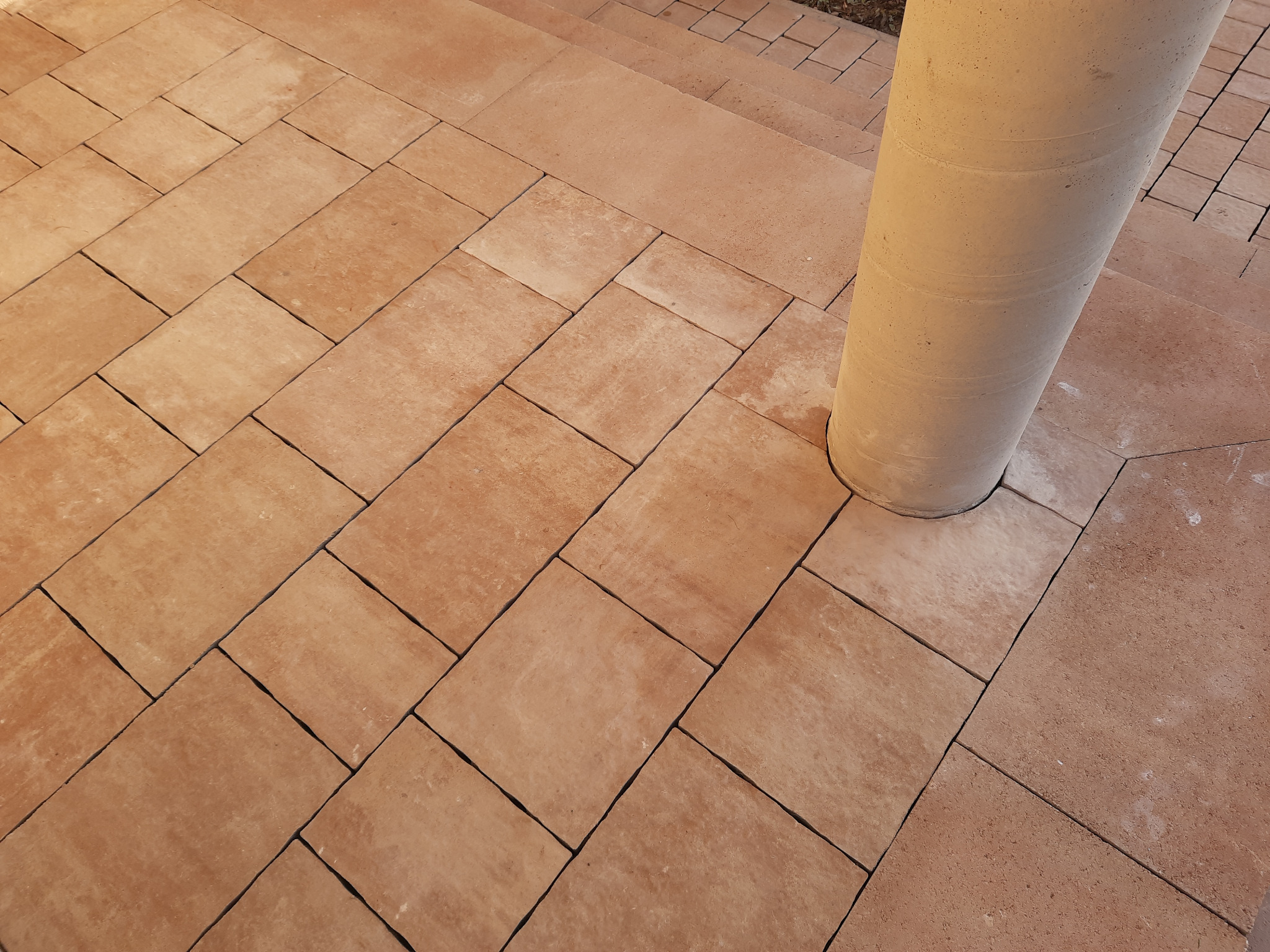 Ułożona, pomarańczowo-brązowa kostka brukowa wokół betonowego słupa, prowadząca do schodów.