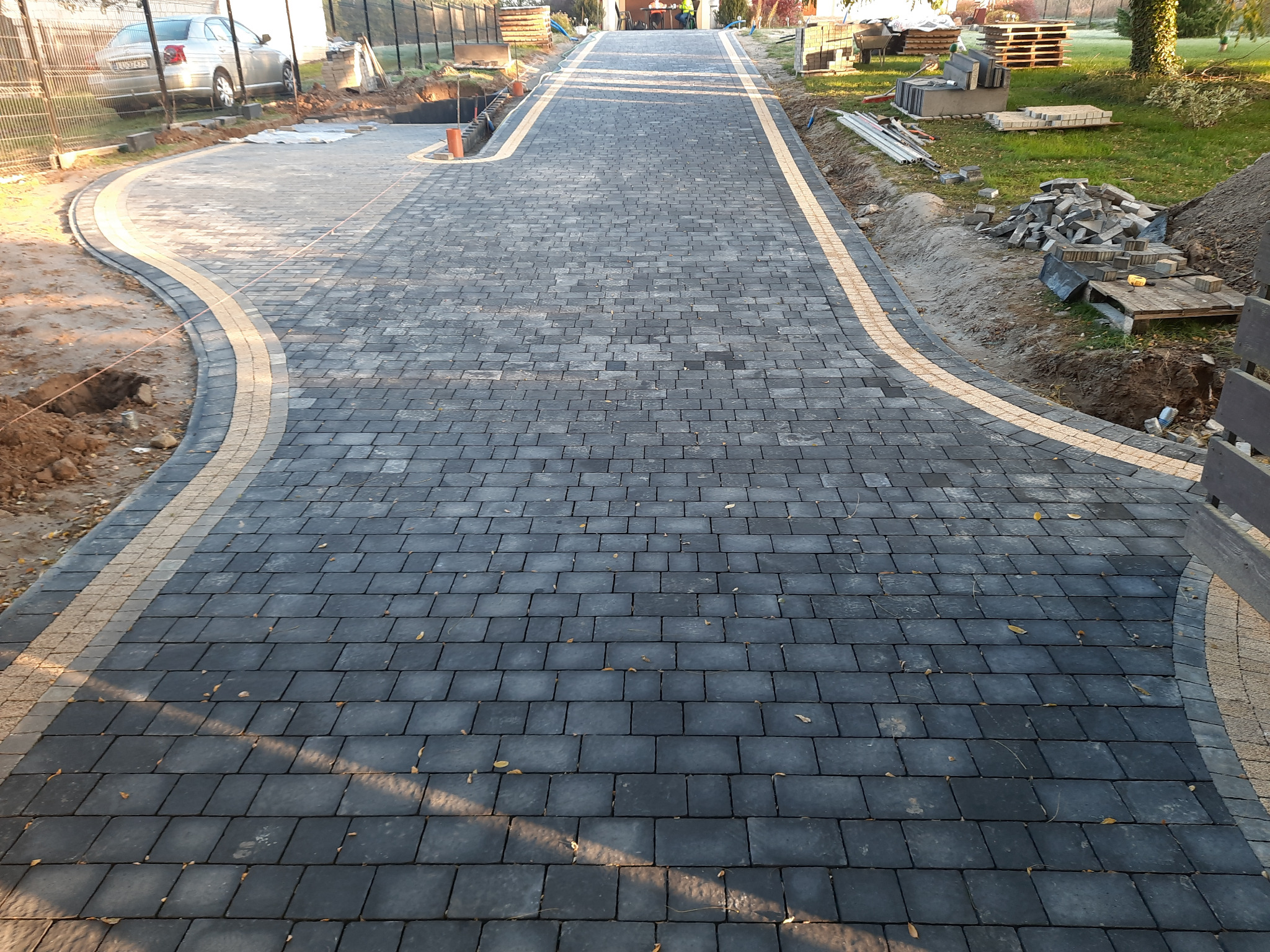 Nowo ułożona, ciemnoszara kostka brukowa z jasną obwódką, tworząca ścieżkę w ogrodzie, widoczna w perspektywie z bliska, z elementami placu budowy po bokach.