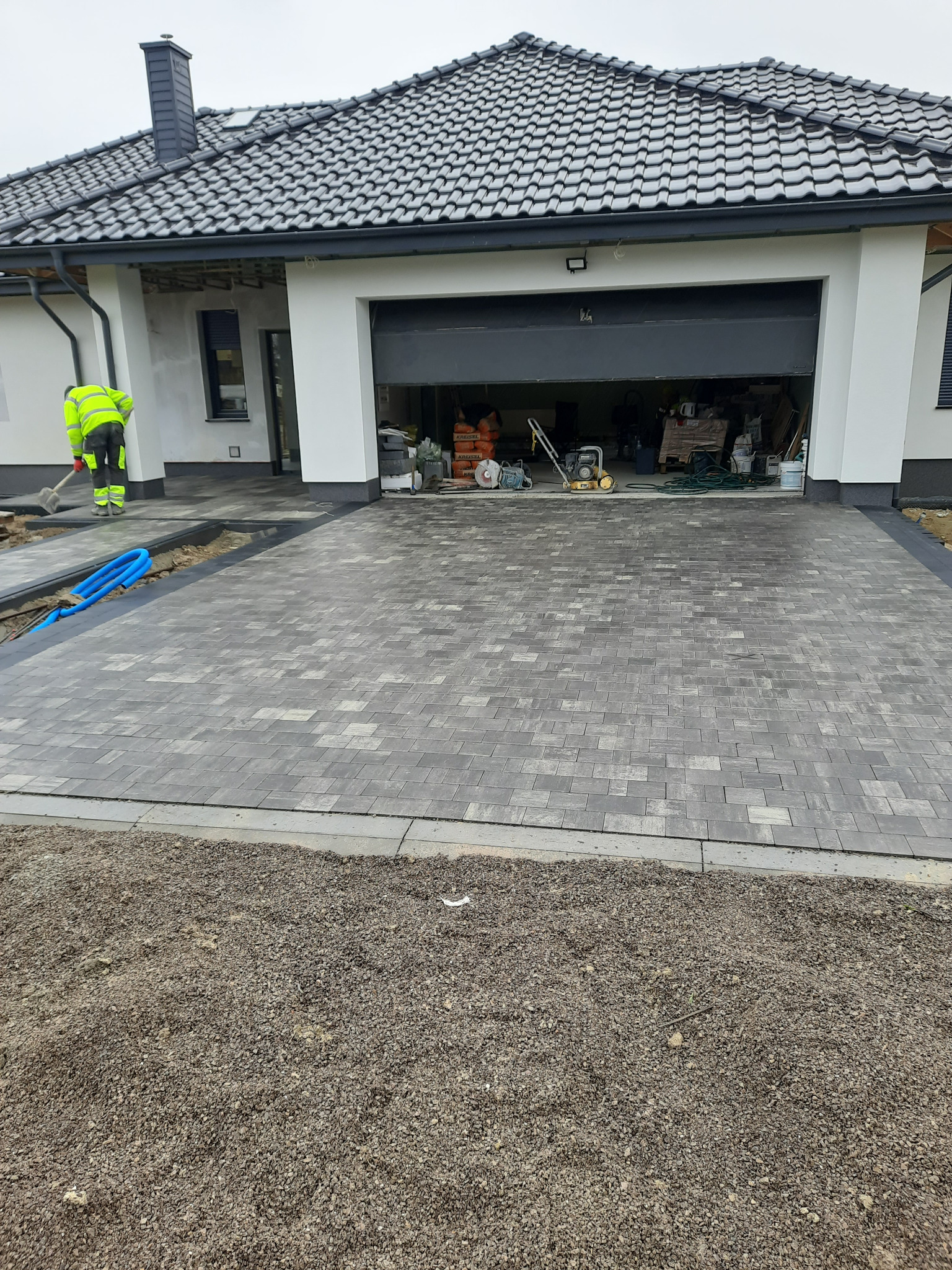 Układanie szarej kostki brukowej na podjeździe do garażu, widoczny pracownik w odblaskowej kamizelce i niebieskie rury, z tyłu otwarty garaż z narzędziami.