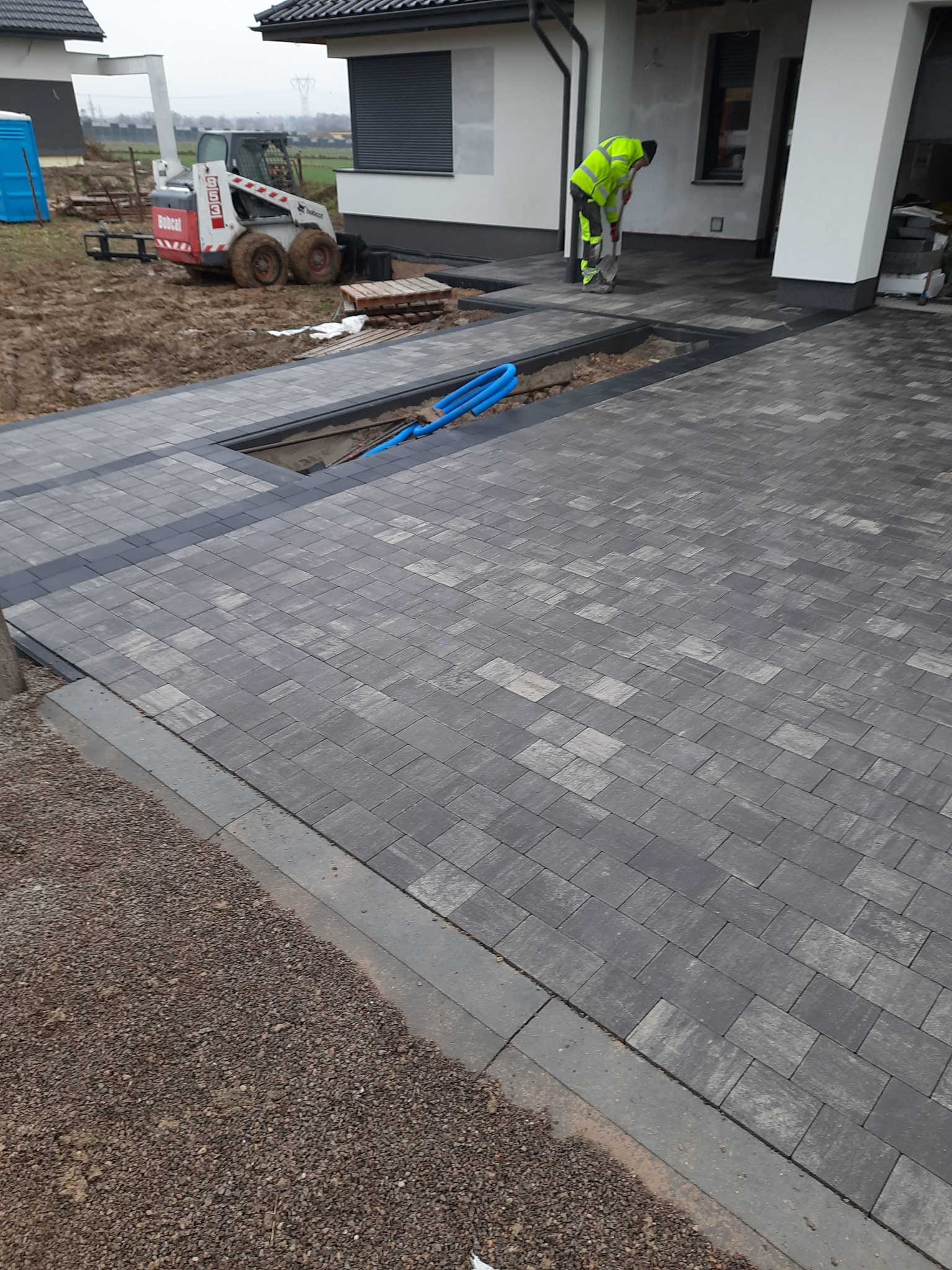 Ułożona szara kostka brukowa przed domem, z widocznym fragmentem wykopu z niebieskimi rurami, pracownikiem w odblaskowej kamizelce i małą koparką Bobcat w tle.