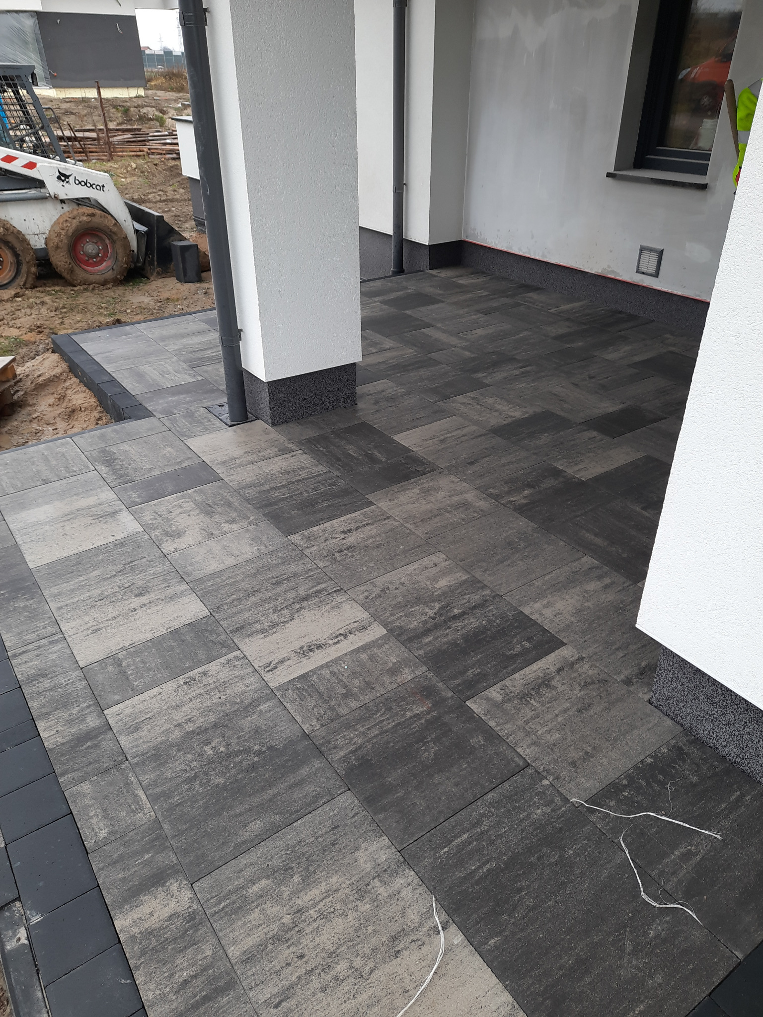 Ułożona, szara kostka brukowa na tarasie przy domu z widoczną miniładowarką Bobcat w tle.