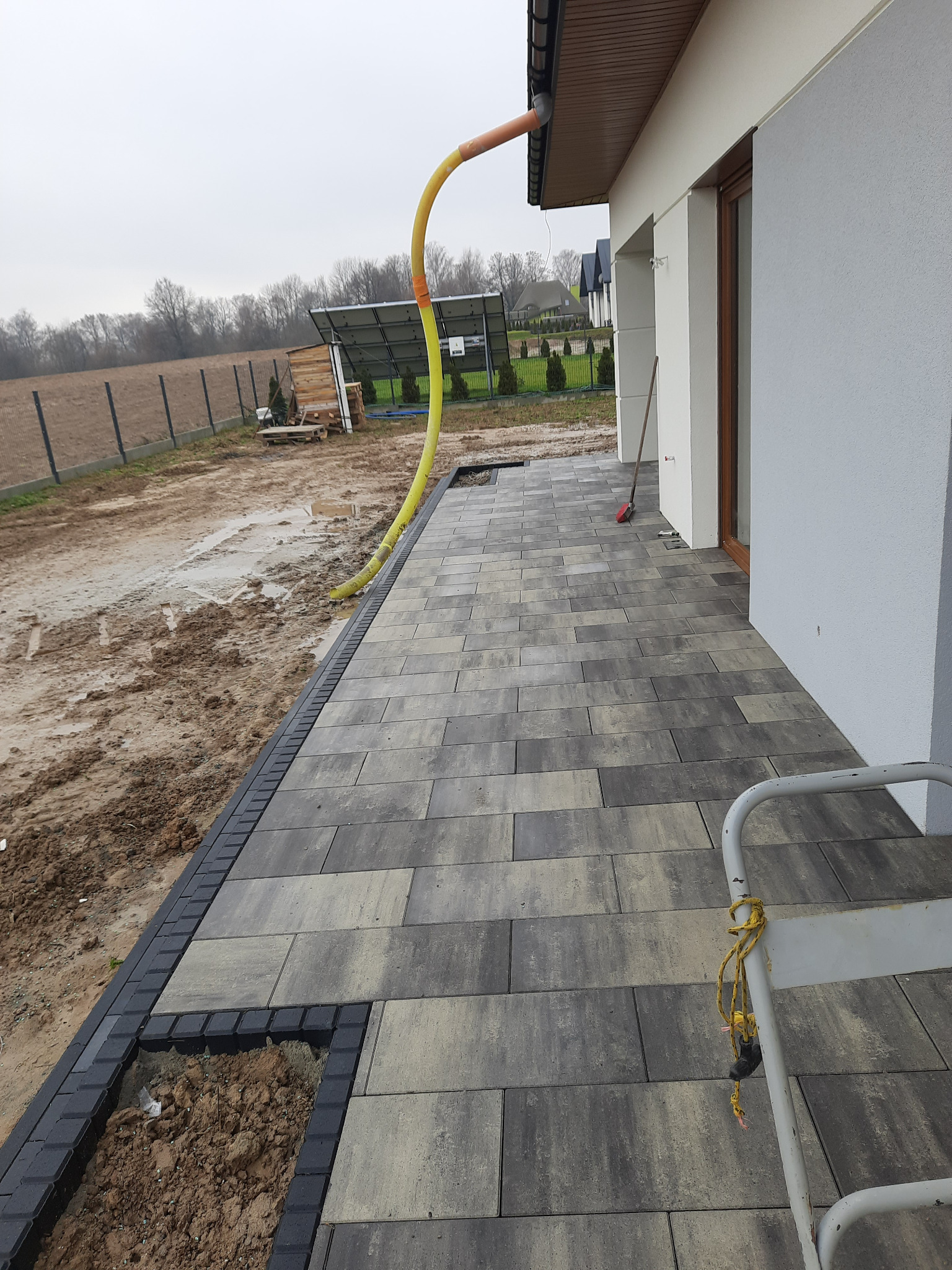 Ułożona kostka brukowa w odcieniach szarości i beżu przy białym budynku, z widoczną żółtą rurą spustową i fragmentem niezakończonej obudowy z kostki przy ziemi.