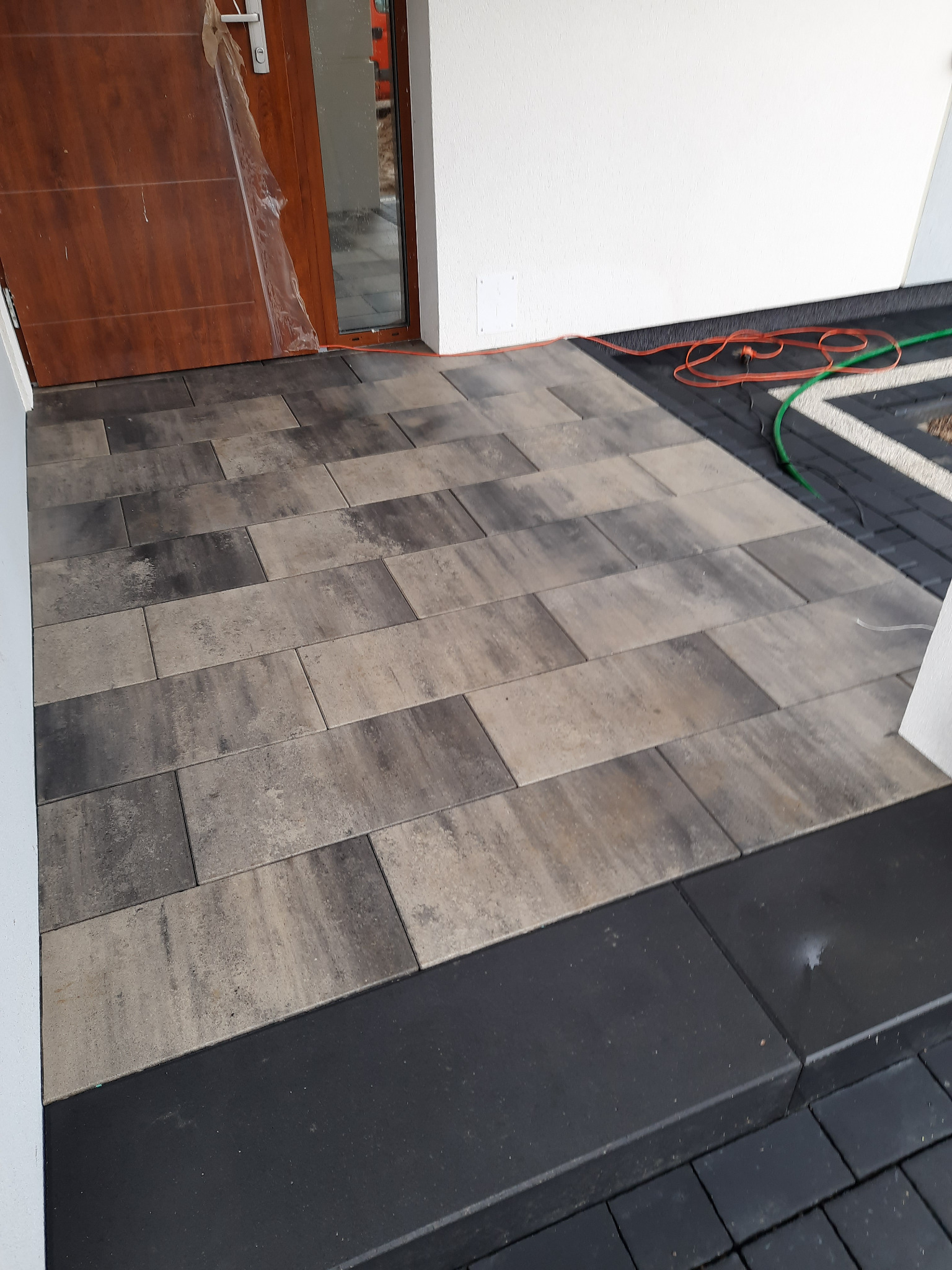 Ułożona kostka brukowa w odcieniach szarości i beżu przed wejściem do domu, z widocznymi przewodami elektrycznymi i częściowo zakrytymi folią drzwiami.