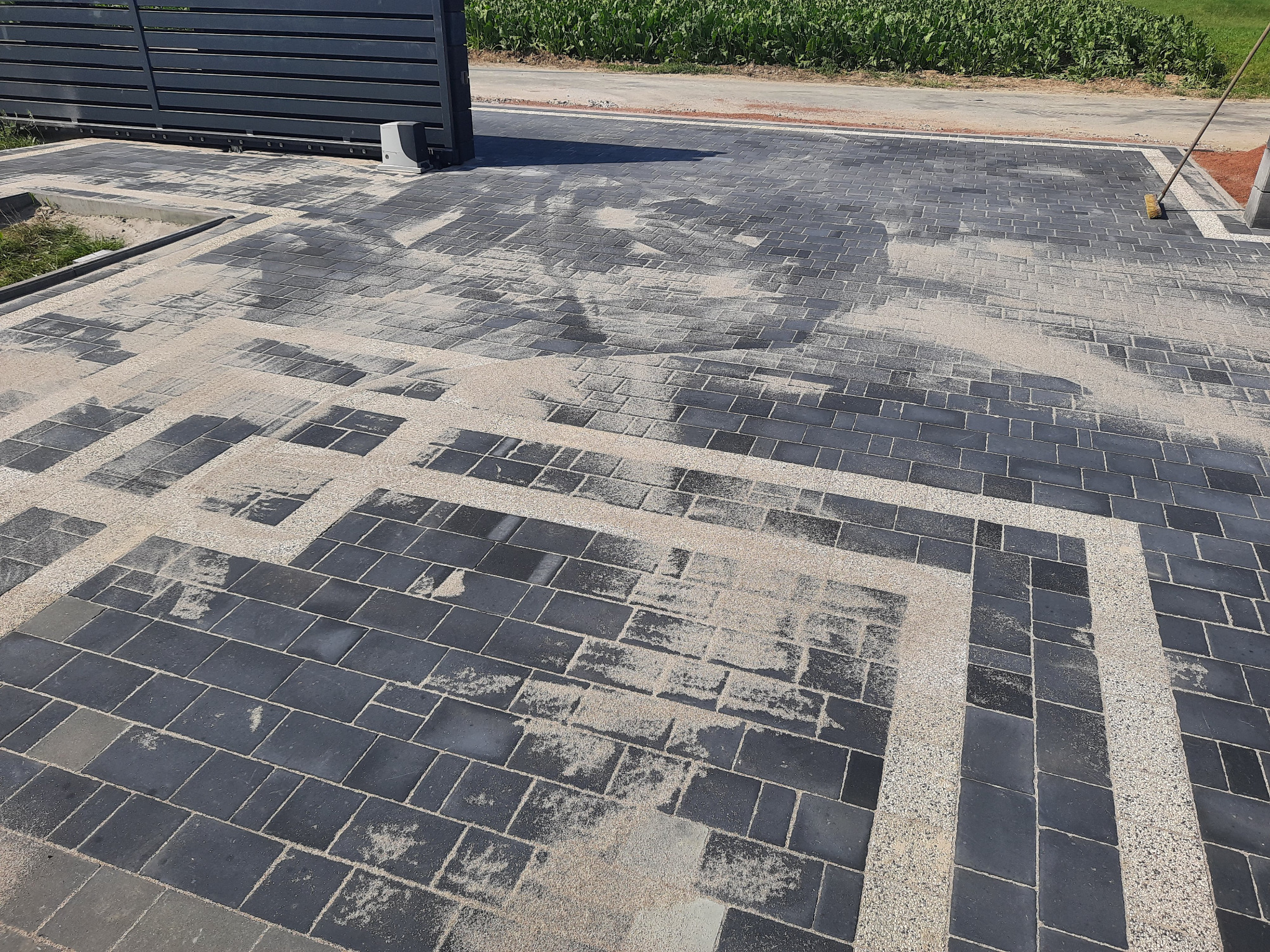 Nowo ułożona, czarna kostka brukowa z jasnymi akcentami wzdłuż bramy wjazdowej, zasypana piaskiem, widoczna szczotka do zamiatania.