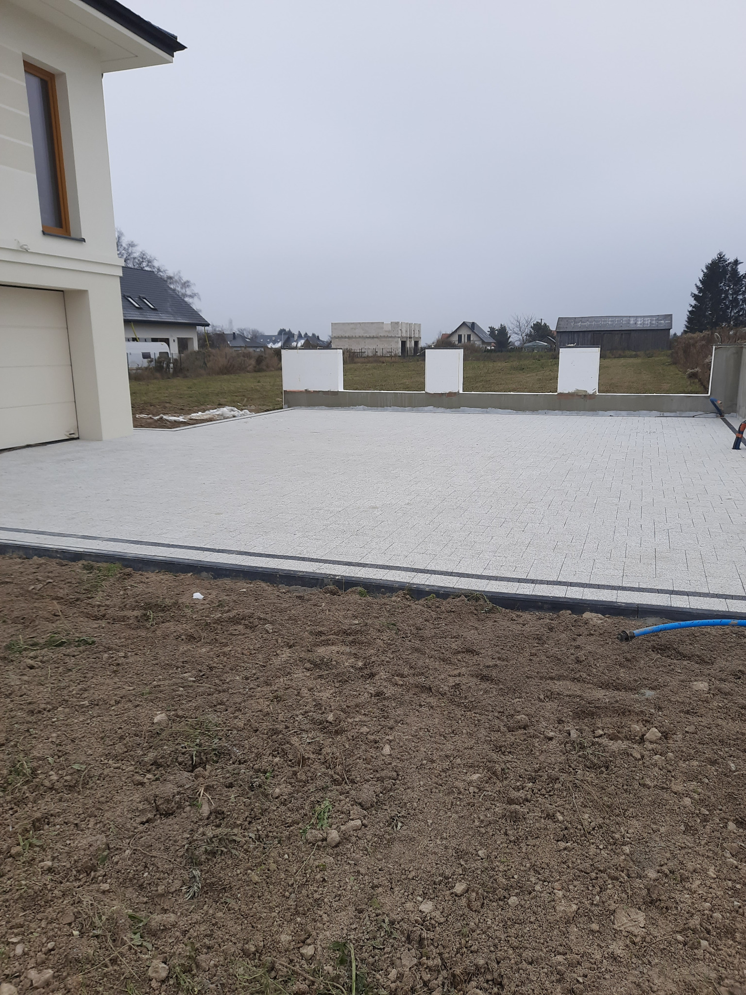 Jasnoszara kostka brukowa ułożona na podjeździe przed nowym domem, widoczne przygotowane obrzeża i częściowo odsłonięta ziemia.