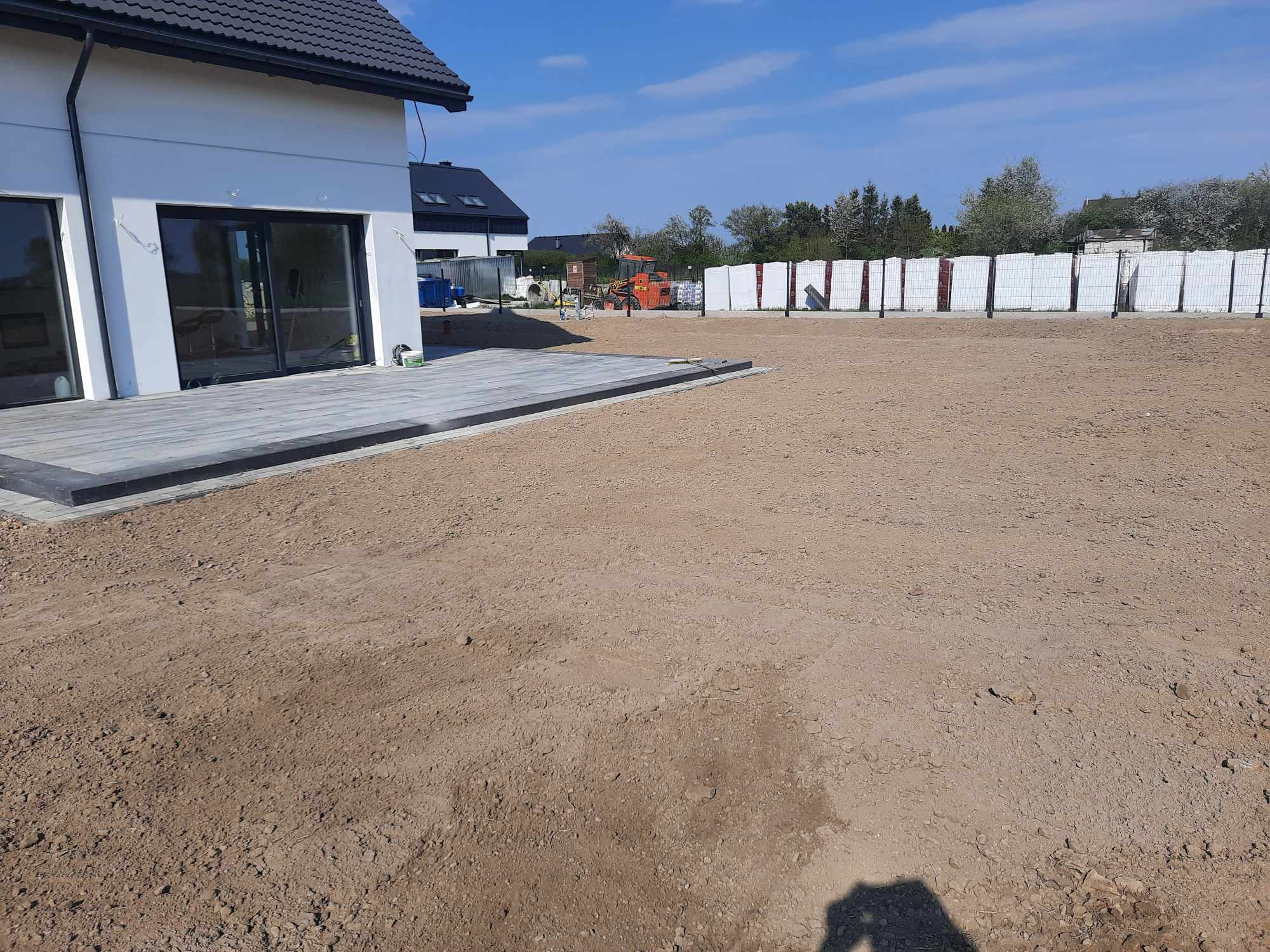 Przygotowany teren pod budowę z widoczną podbudową pod taras z szarych płyt, w tle palety z materiałami budowlanymi i maszyna budowlana.