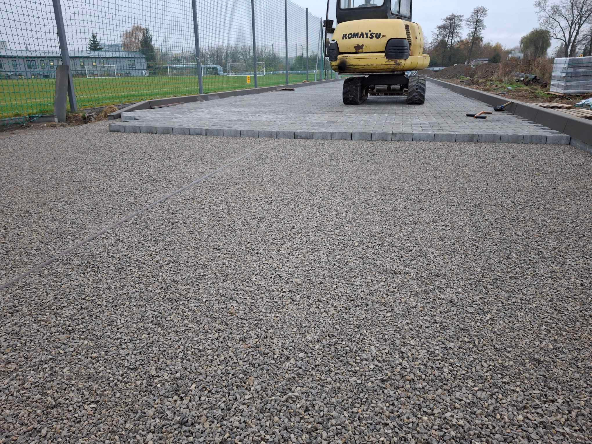 Przygotowanie podłoża pod kostkę brukową: warstwa tłucznia, obrzeża betonowe i częściowo ułożona nawierzchnia z szarej kostki, widoczna mini koparka Komatsu oraz narzędzia.