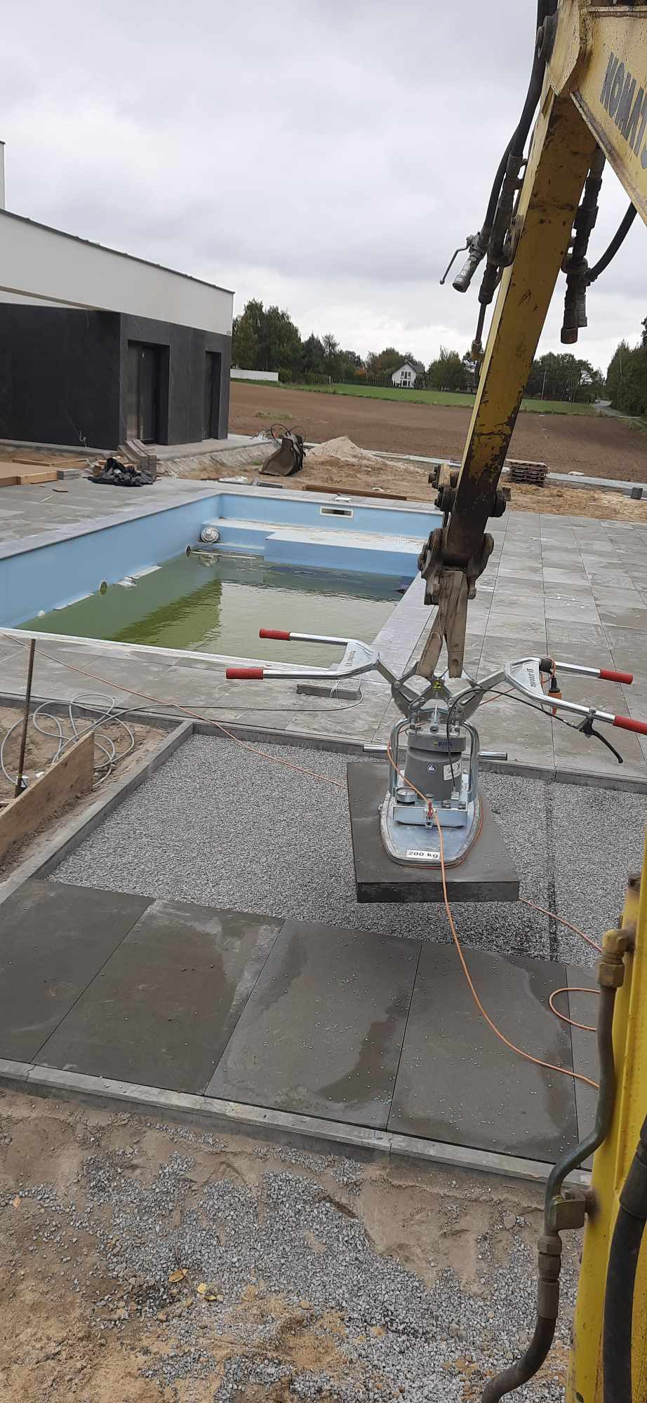 Żuraw z przyssawką umieszcza dużą płytę chodnikową na podsypce z kruszywa, obok basenu w trakcie budowy w Lublinie.