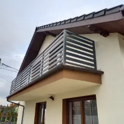 Nowoczesny balkon z poziomymi, ciemnymi panelami na tle białej elewacji domu z brązowym dachem i rynnami.