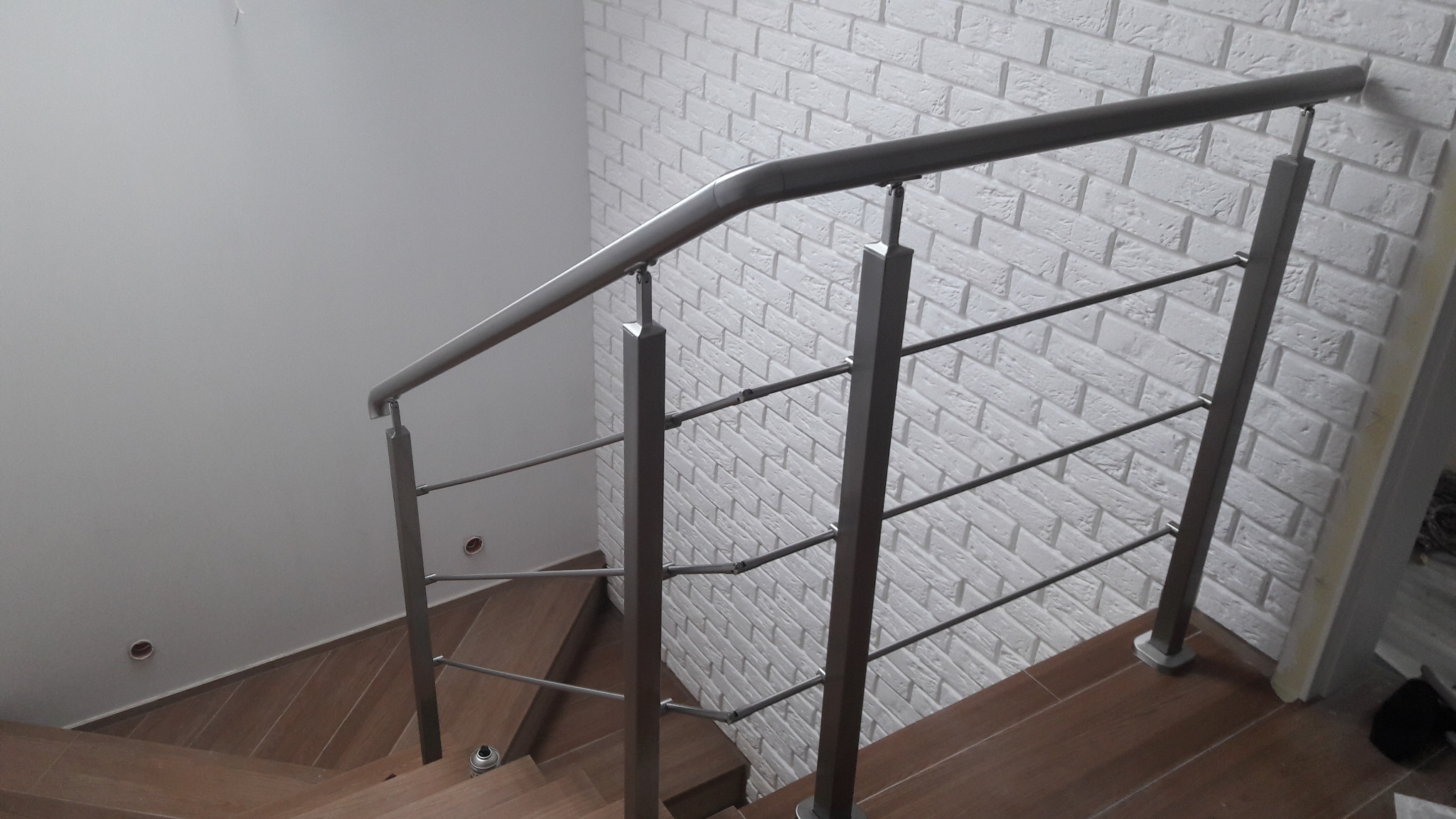 Nowoczesne schody z drewnianymi stopniami i metalową balustradą z poziomymi prętami na tle ściany z białej cegły.