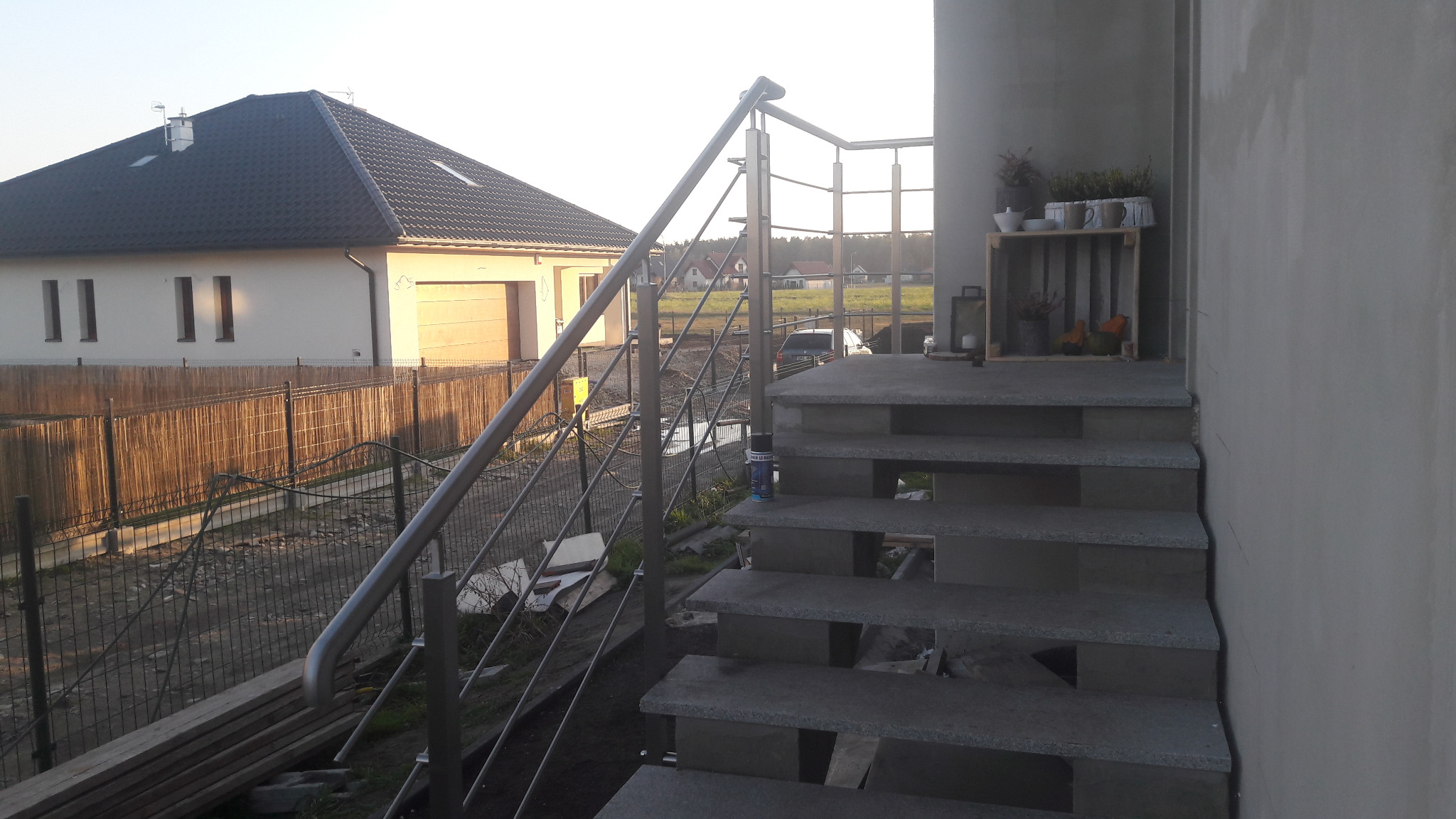 Zewnętrzne schody z szarego kamienia z metalową balustradą, widok na dom jednorodzinny i ogrodzenie z siatki.