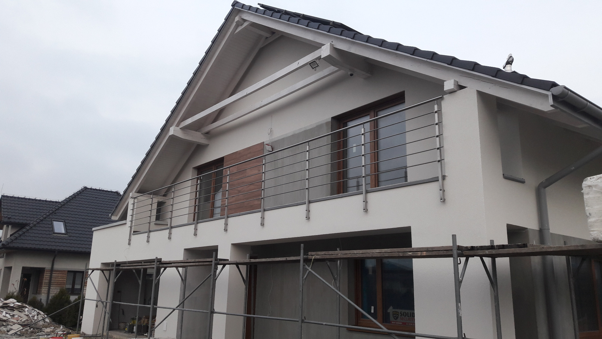Nowoczesny dom z balkonem zabezpieczonym prostą, poziomą konstrukcją ze stali, widok z zewnątrz na elewację w trakcie prac wykończeniowych, rusztowania w dolnej części kadru.