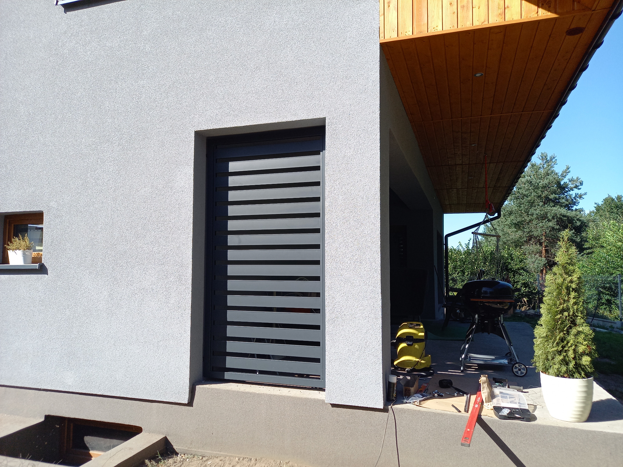 Szara, pozioma żaluzja zewnętrzna zamontowana w elewacji domu, widoczne narzędzia montażowe i grill w tle.
