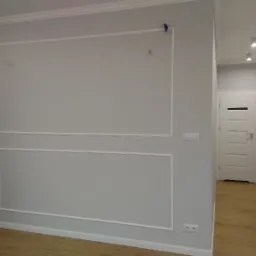 Szara ściana z dekoracyjnymi białymi listwami tworzącymi prostokątne ramy, ciemne zasłony po lewej stronie, widok na korytarz z białymi drzwiami i oświetleniem punktowym.