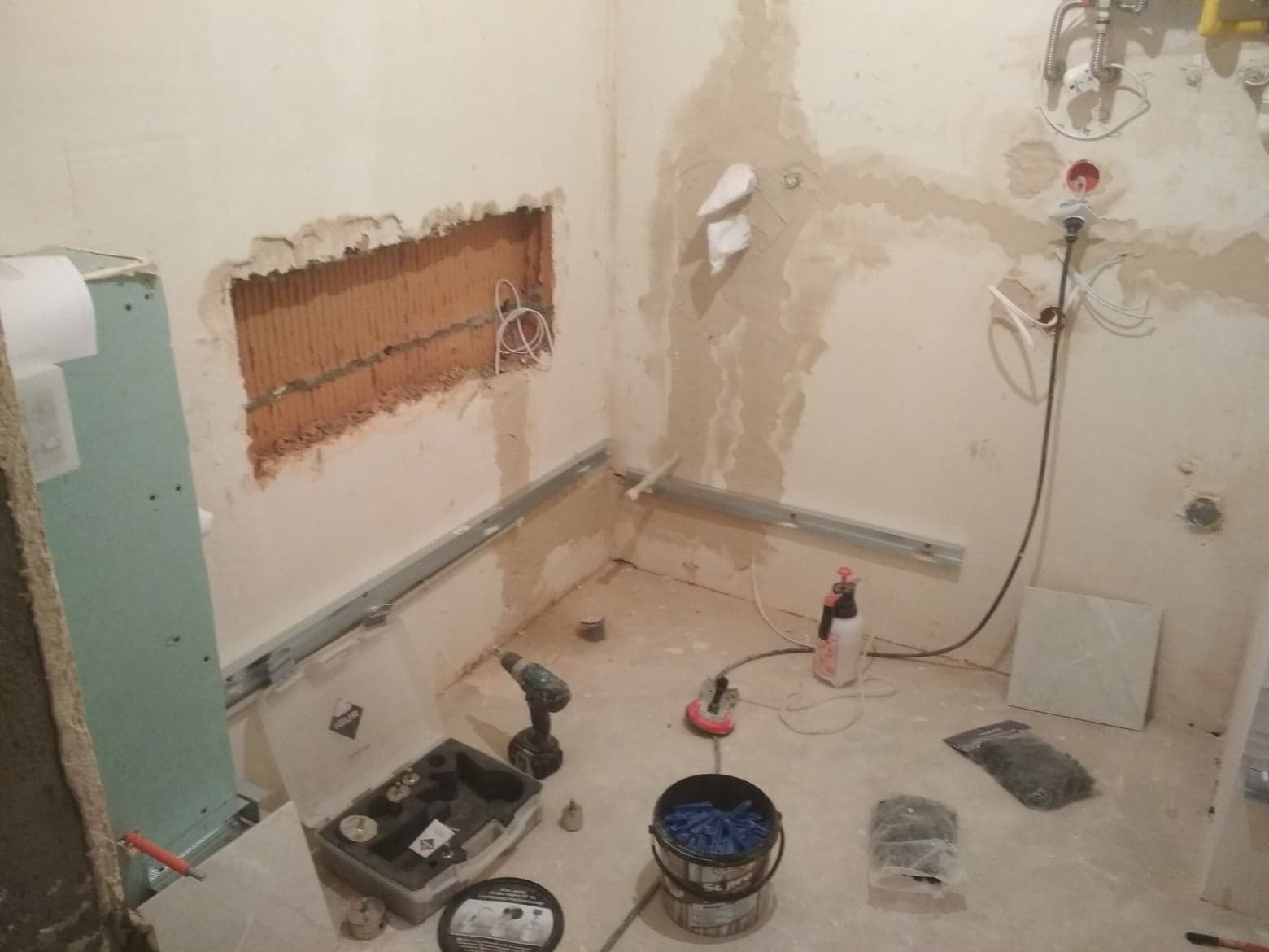 Łazienka w trakcie prac przygotowawczych do położenia płytek: odsłonięte przewody, metalowe profile ścienne, narzędzia glazurnicze, wiercone otwory w ścianach.