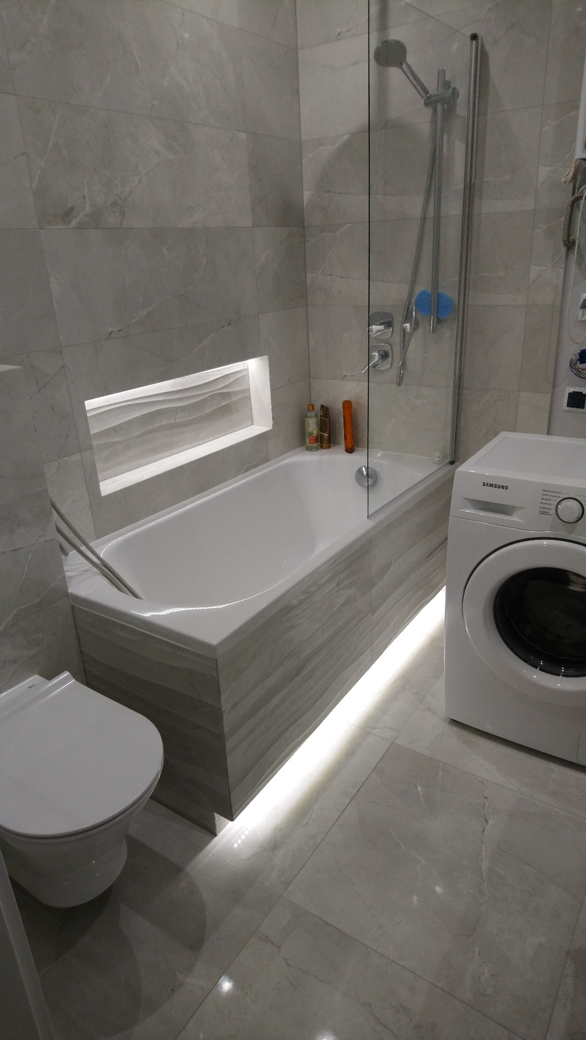 Wykończona łazienka w szarościach z podświetlaną wanną, szklanym parawanem nawannowym, toaletą i pralką Samsung.