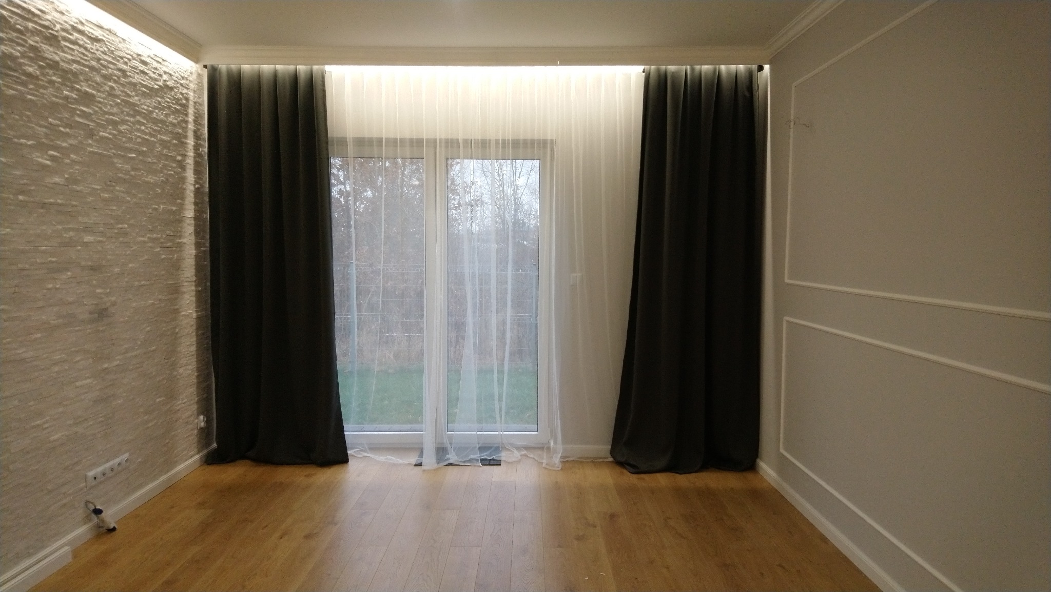 Pokój z drewnianą podłogą, ścianą z dekoracyjną fakturą kamienia, ścianą z dekoracyjnymi listwami, oknem z białymi firanami i ciemnymi zasłonami