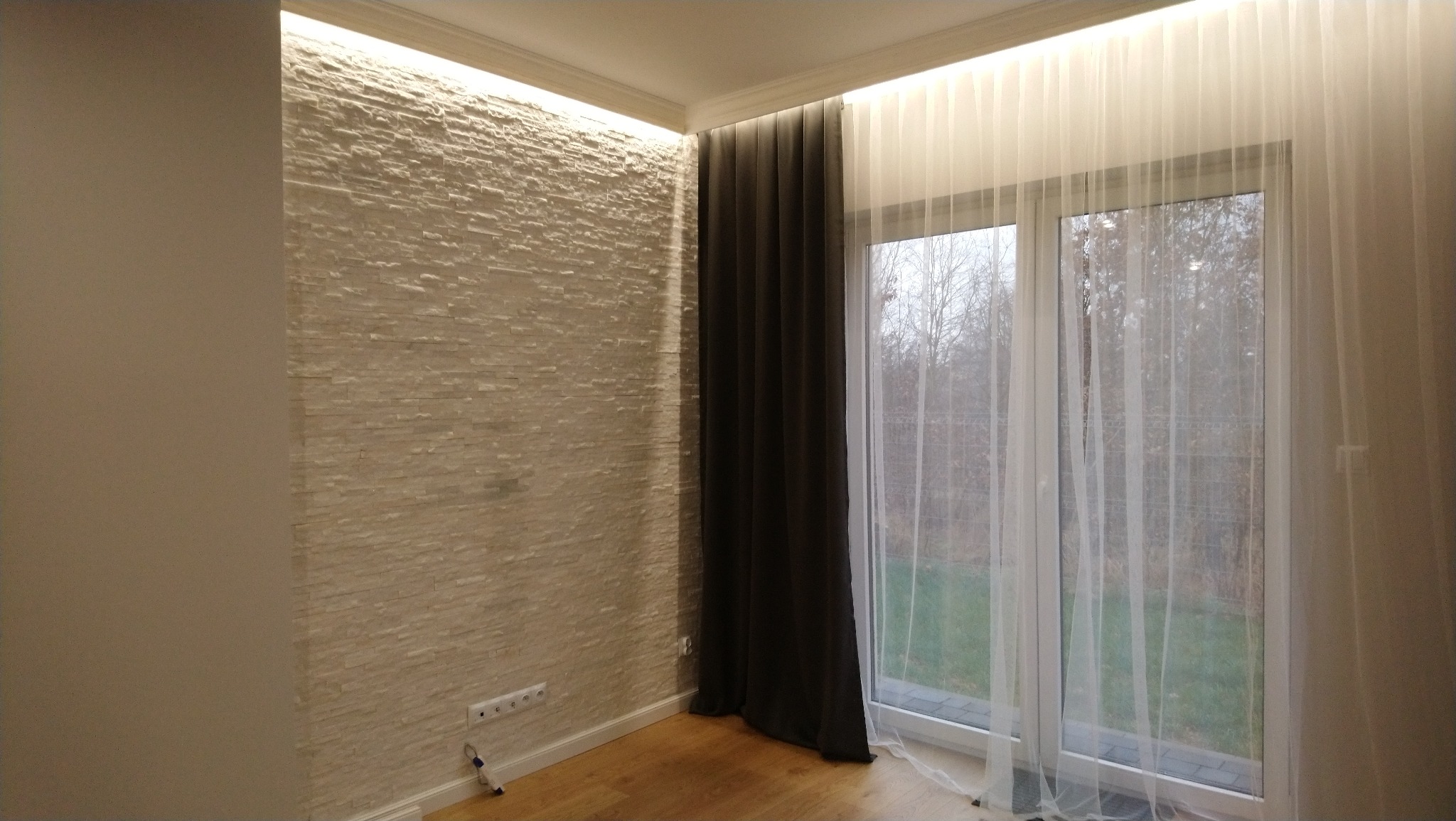 Pokój z dekoracyjną ścianą z kamienia, oświetleniem LED podsufitowym, oknem z białymi i grafitowymi zasłonami oraz drewnianą podłogą.