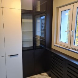 UNIFOX Grzegorz Lis - Nowoczesna zabudowa kuchenna z białymi frontami i czarną witryną z przeszklonymi półkami, marmurowa podłoga, widoczny grzejnik zabudowany w czarnej obudowie pod oknem.
