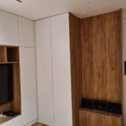 Nowoczesna szafa wnękowa z białymi frontami i drewnianym panelem ściennym, z widocznymi przewodami oświetleniowymi na suficie i podłogą ułożoną w jodełkę.