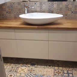 Łazienka z drewnianym blatem, białą umywalką nablatową i szafką pod umywalką z białymi frontami, widoczna przez otwarte drzwi, z wzorzystą glazurą na ścianie i podłodze.