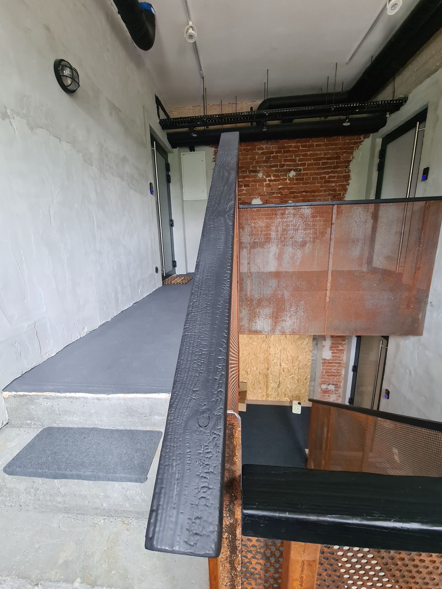 Betonowe schody z czarną drewnianą balustradą i metalową ażurową barierką w loftowym wnętrzu z surowymi ścianami z cegły i betonową posadzką.