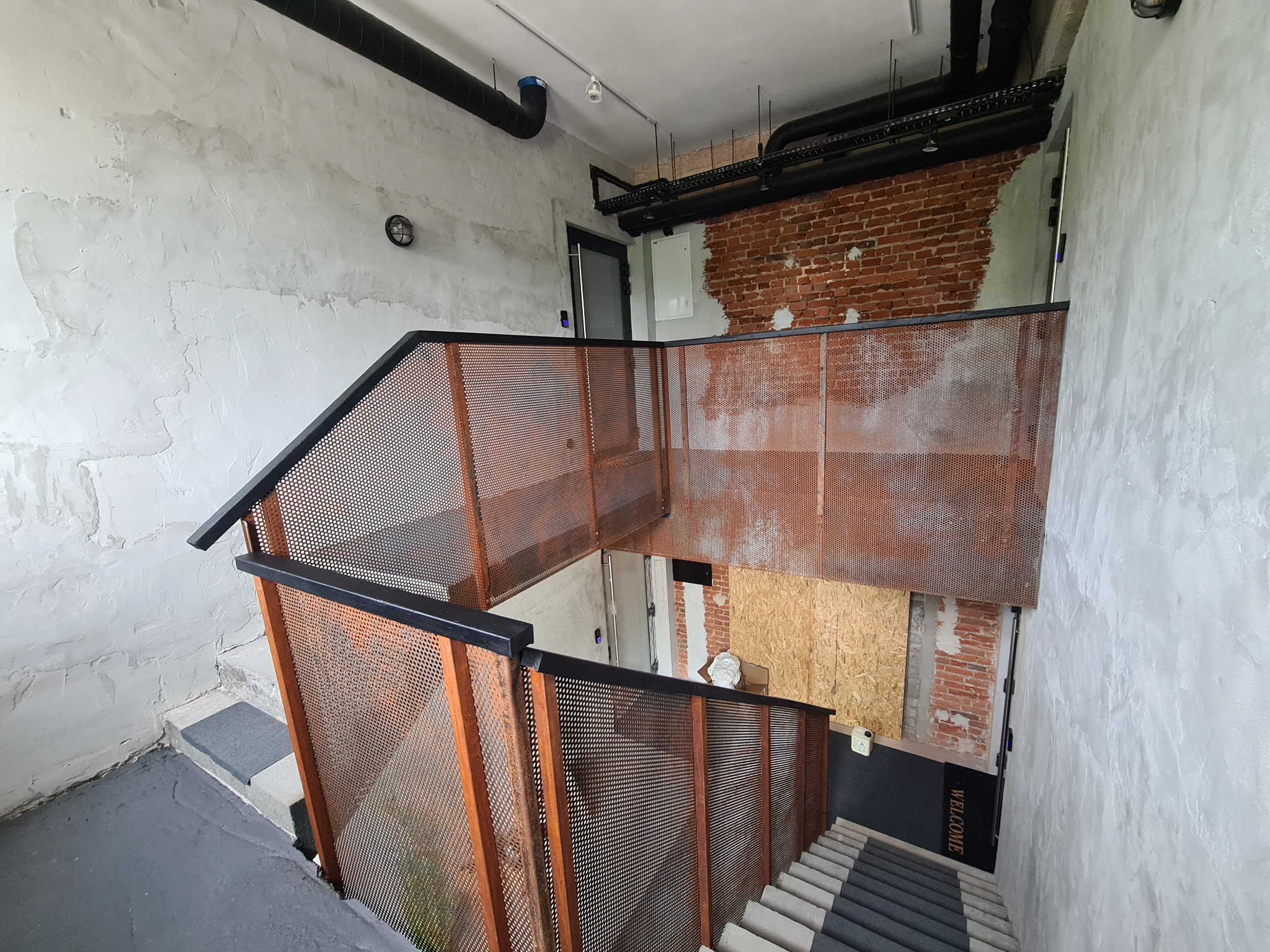 Schody z metalową ażurową balustradą w stylu loftowym, widok z góry, surowe ściany z fragmentami cegły, widoczne instalacje pod sufitem.