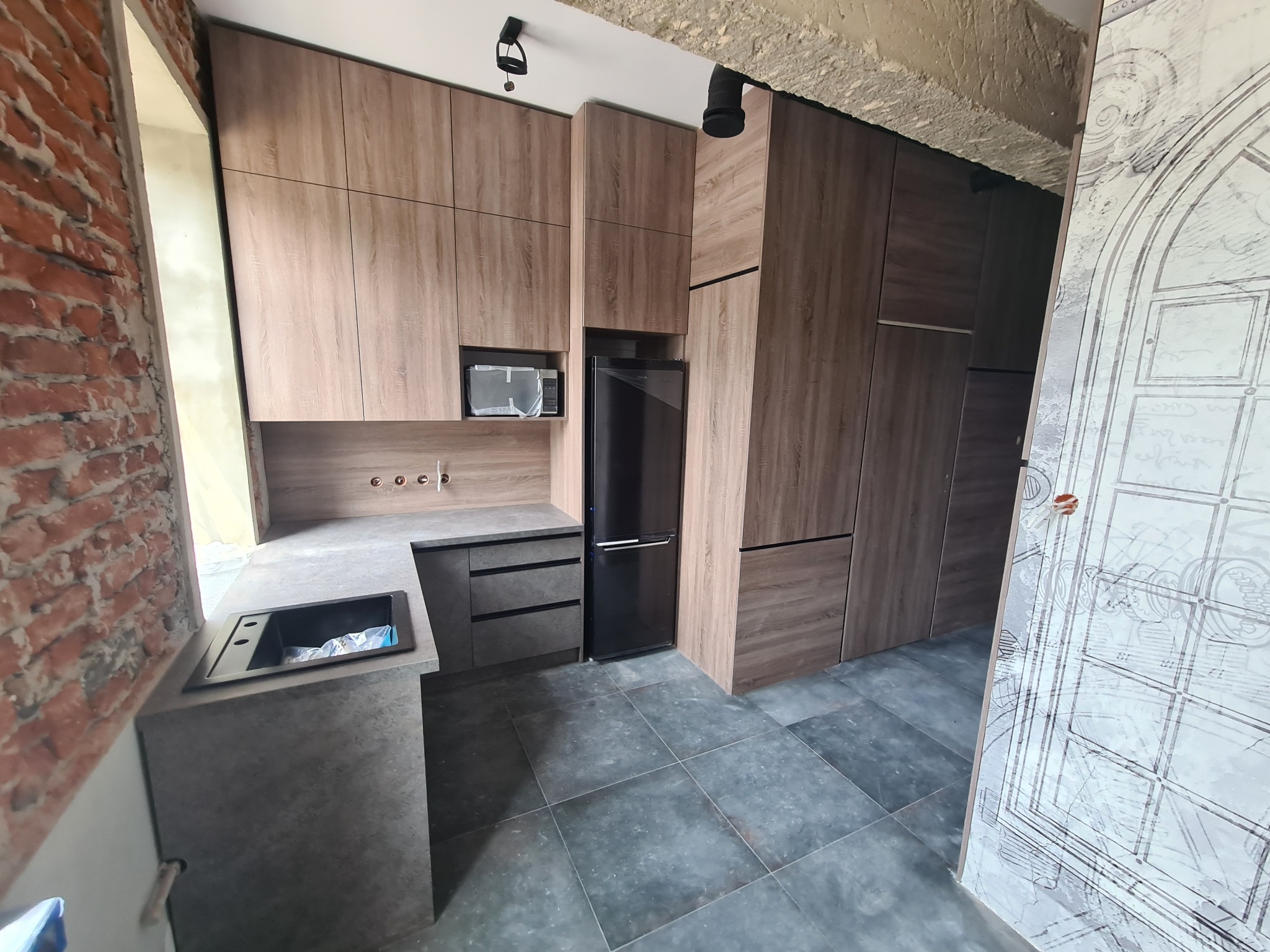 Nowoczesna zabudowa kuchenna z drewnopodobnymi frontami, szarym blatem i czarną lodówką w surowym wnętrzu z ceglaną ścianą i betonowym sufitem. Widoczne instalacje elektryczne i grafika na drzwiach.