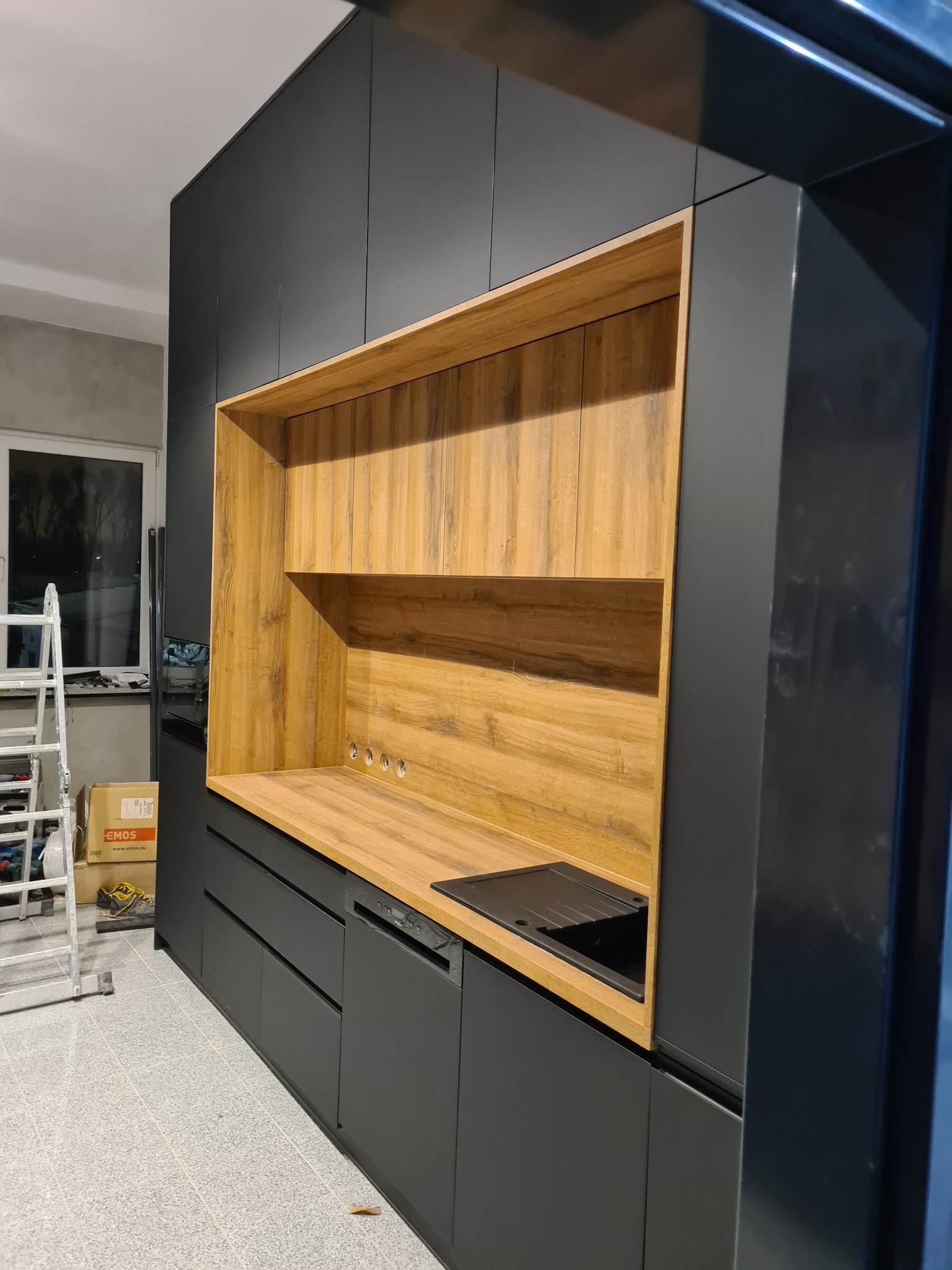 Ciemnoszara zabudowa kuchenna z drewnianym blatem i otwartymi półkami z drewnianym wykończeniem, widoczny zlewozmywak i drabina w tle.