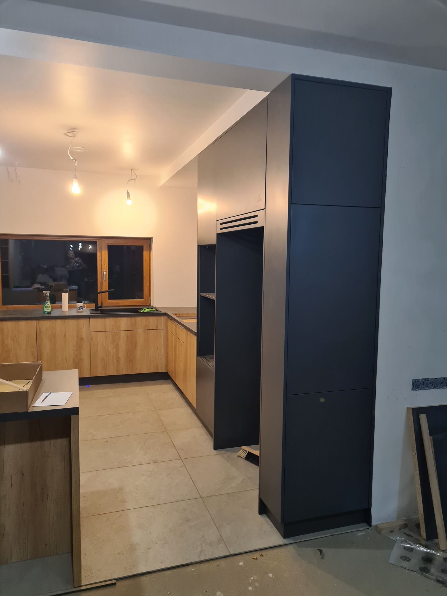 Częściowo zamontowana kuchnia z drewnianymi frontami i ciemnoszarymi wysokimi szafkami, widoczne okno, blat i podłoga z płytek, instalacja w trakcie.