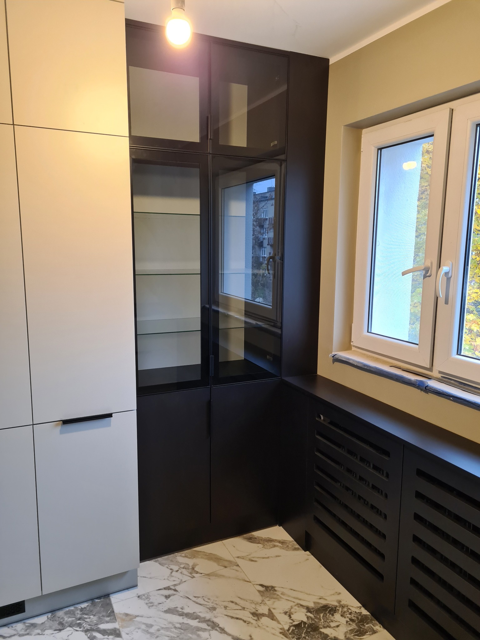 Nowoczesna zabudowa kuchenna z białymi frontami i czarną witryną z przeszklonymi półkami, marmurowa podłoga, widoczny grzejnik zabudowany w czarnej obudowie pod oknem.