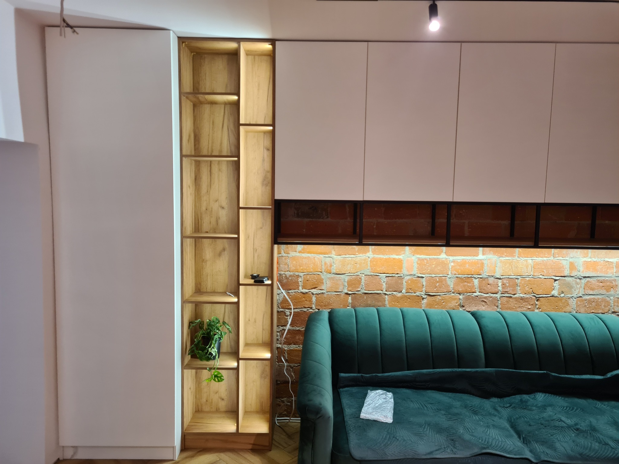 Nowoczesny salon z białą zabudową meblową, drewnianym regałem z podświetleniem LED, zieloną sofą i ścianą z cegły.