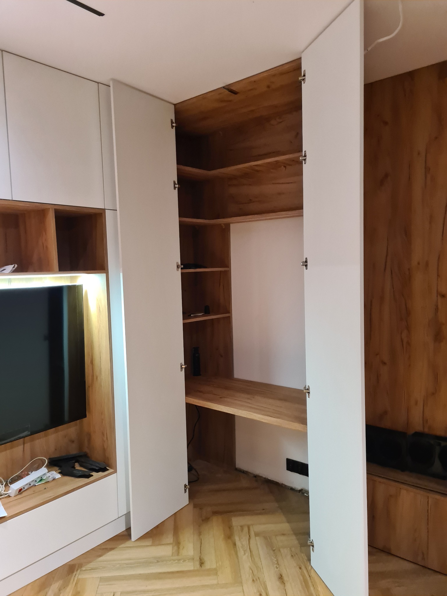 Biała szafa wnękowa z otwartymi drzwiami, ukazująca drewniane półki i biurko wewnątrz, wkomponowana w zabudowę ściany z elementami drewnopodobnymi.