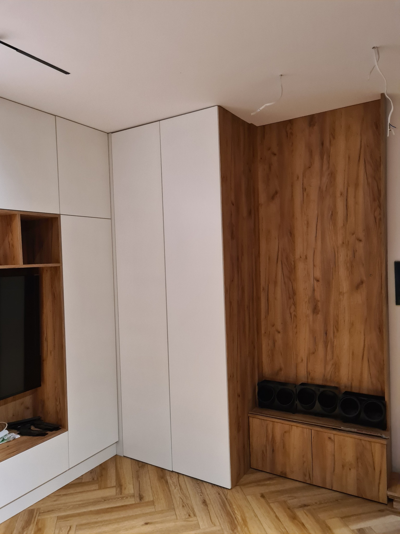Nowoczesna szafa wnękowa z białymi frontami i drewnianym panelem ściennym, z widocznymi przewodami oświetleniowymi na suficie i podłogą ułożoną w jodełkę.