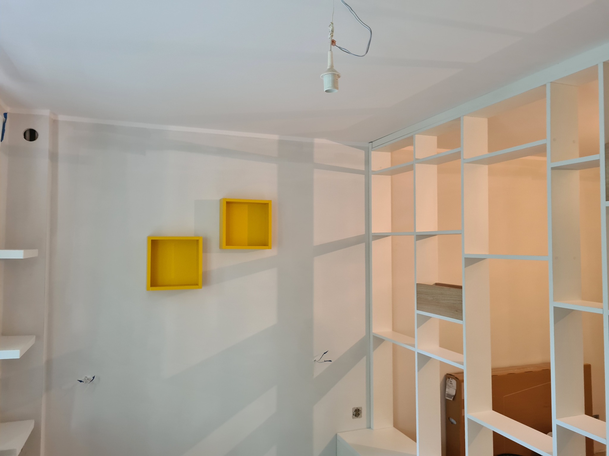 Wnętrze w trakcie wykańczania: biała, modułowa konstrukcja regałowa na ścianie, dwa żółte, kwadratowe elementy dekoracyjne, widoczne kable i puszka elektryczna, surowa żarówka bez klosza.