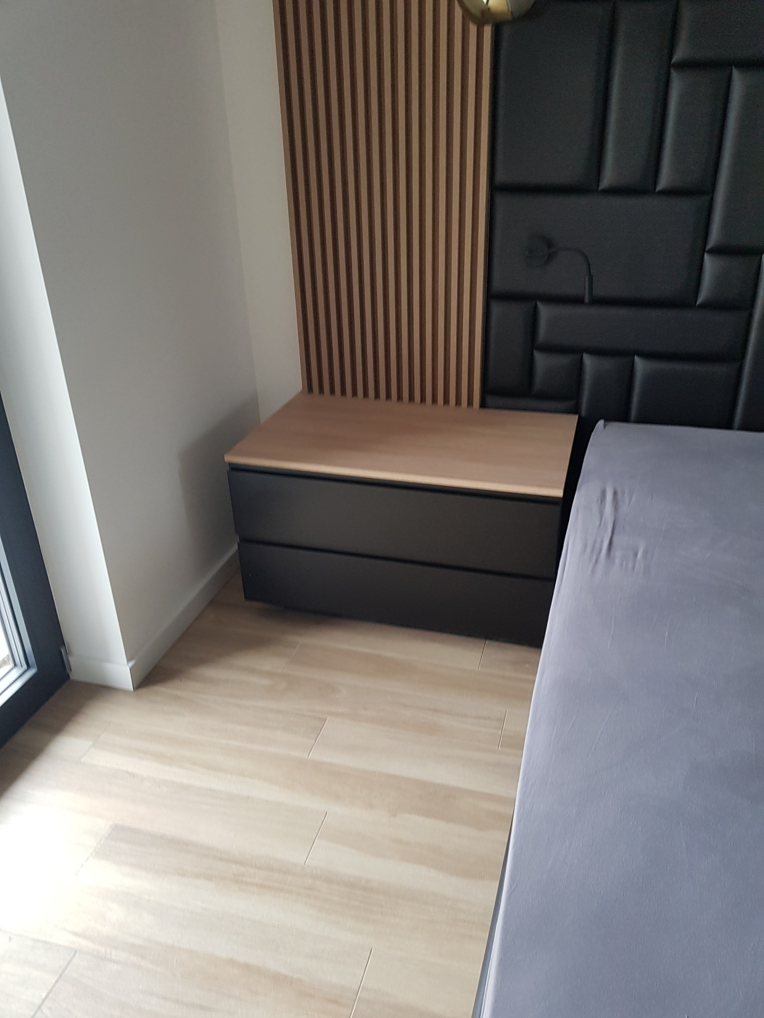 Nowoczesna szafka nocna z dwoma szufladami, z drewnianym blatem, umieszczona przy ścianie z drewnianymi lamelami i tapicerowanym panelem zagłówka, obok łóżka z szarą pościelą.