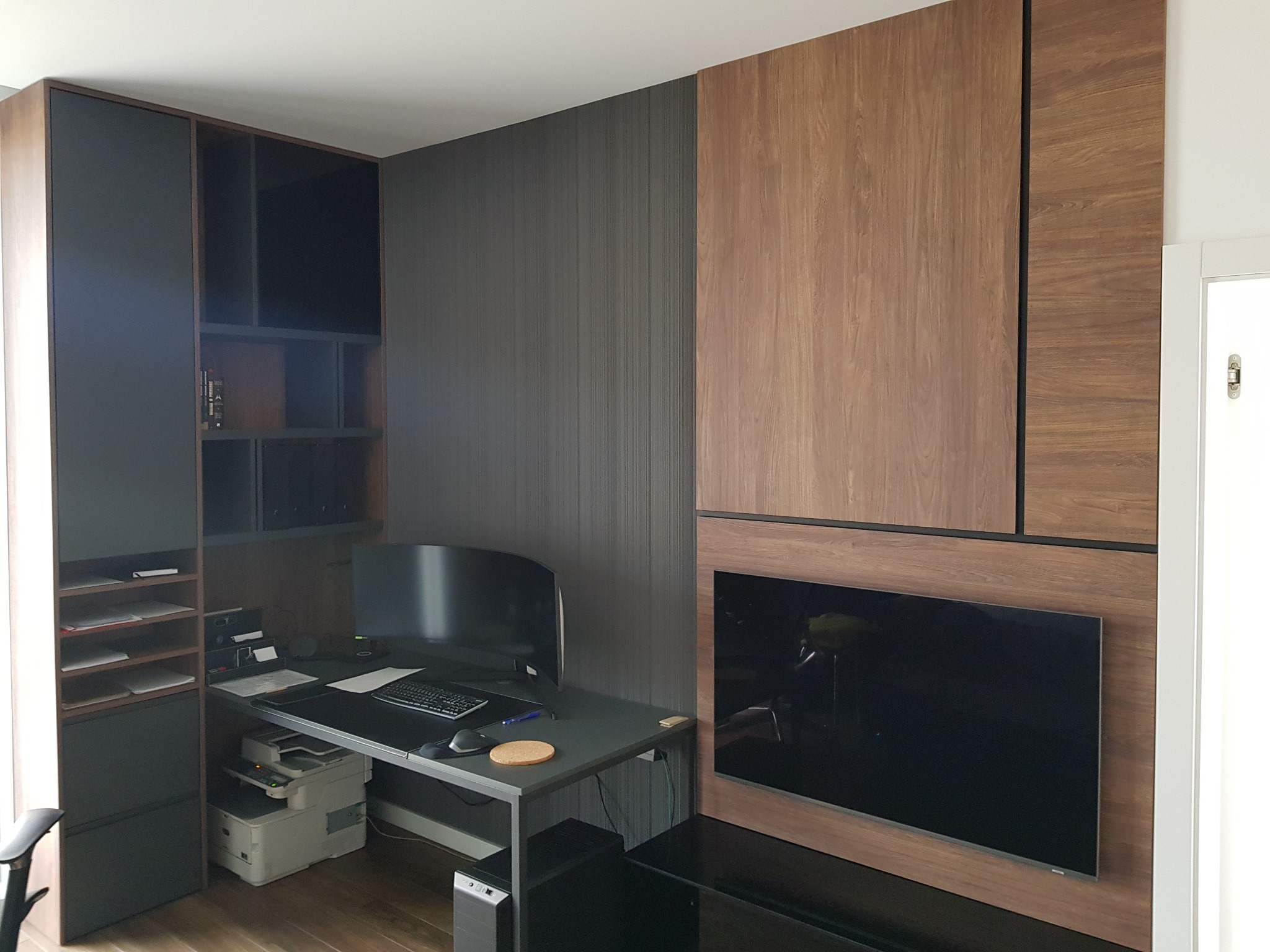 Nowoczesne biuro w domu z zabudową meblową: szafki z drewnianym fornirem i czarnymi elementami, biurko z komputerem i drukarką, telewizor wkomponowany w ścianę.