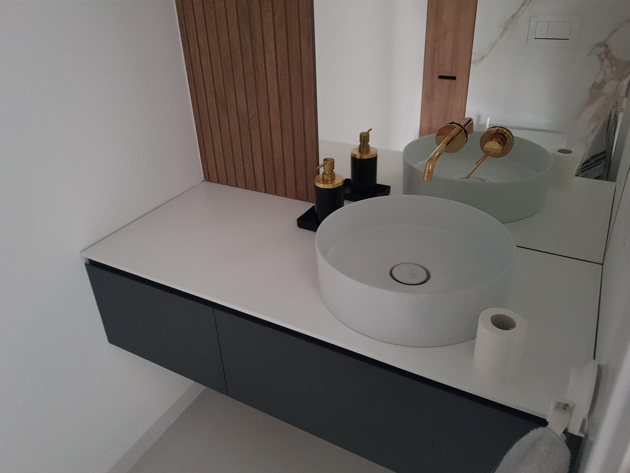 Nowoczesna łazienka z okrągłą, białą umywalką nablatową, złotą armaturą i szafką pod umywalką w kolorze antracytowym, uzupełniona dozownikami mydła i dekoracyjną ścianką z drewnianych paneli.