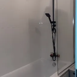 Nowoczesna łazienka z białą wanną obudowaną szarymi panelami, czarną armaturą prysznicową z deszczownicą i szklanym parawanem nawannowym.