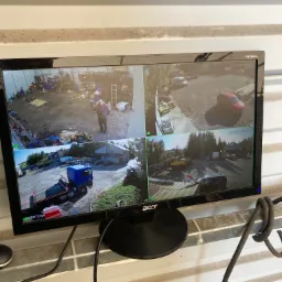 Monitor LCD marki Acer wyświetlający obraz z czterech kamer monitoringu wizyjnego, ukazujących teren zewnętrzny z różnymi pojazdami i budynkami.