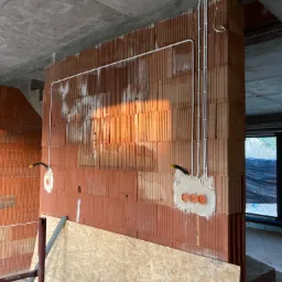 Świeżo położone przewody elektryczne w białych rurkach na ścianie z czerwonej cegły dziurawki, widoczne pomarańczowe puszki instalacyjne, surowy betonowy strop, fragment okna i płyta OSB oparta...